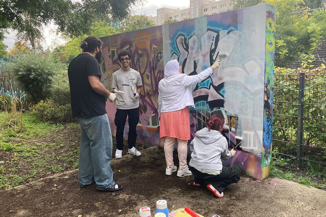 FBS Dresden: Gemeinsames Gestalten einer Graffiti-Wand Mehrere Personen arbeiten gemeinsam an einer bunt bemalten Graffiti-Wand im Freien. Sie tragen Handschuhe und nutzen Farbrollen, um die Wand zu gestalten. Im Hintergrund sind Bäume und ein Wohngebäude zu sehen.
