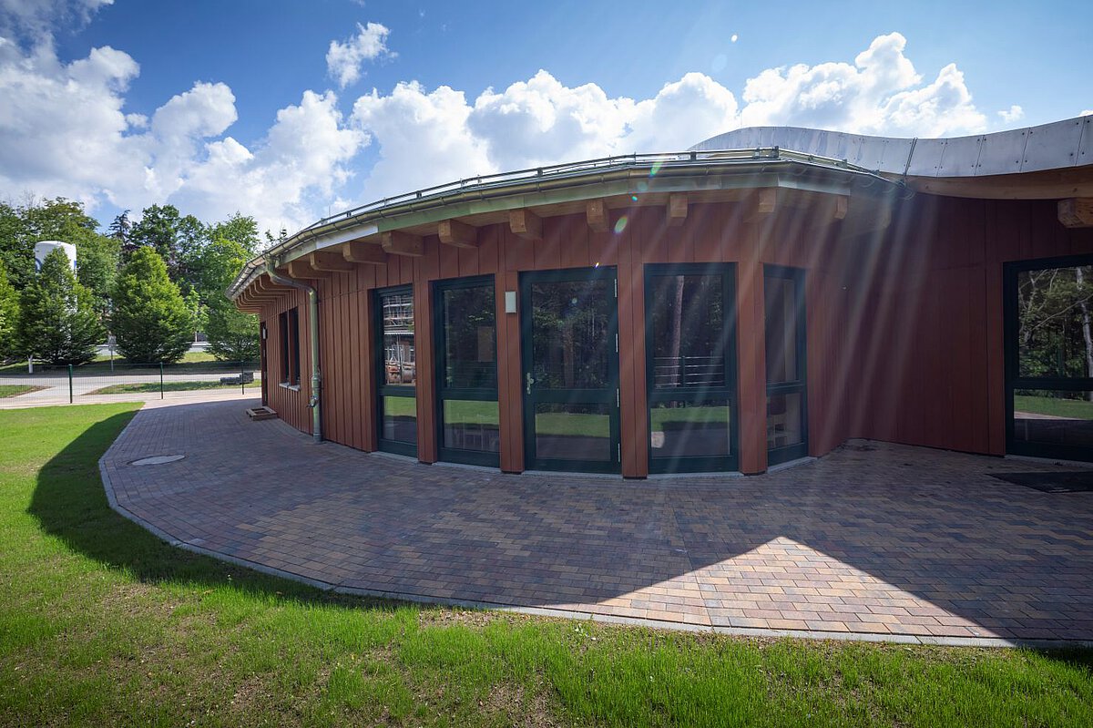 Rückseite des Kindergarten Am Wald- Klinikum- Gebäudes mit großen Fenstern Rückseite eines runden Gebäudes. Die Fassade ist rotbraun und hat große Fenster. Vor dem Gebäude liegt ein gepflasterter Weg, daneben ist grüne Wiese. Im Hintergrund stehen viele Bäume. Der Himmel ist blau mit weißen Wolken, und die Sonne scheint hell.