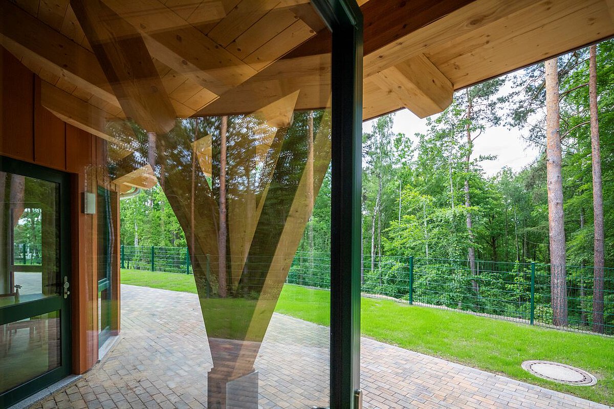 Blick durch Glas auf Pflastersteine und Wald mit Holzstütze Blick aus dem Kindergarten Am Wald- Klinikum Gebäude in Richtung Wald. Im Vordergrund sind große Glasfenster und ein Holzpfeiler, der das Dach stützt. Draußen liegt ein gepflasterter Weg und eine grüne Wiese. Dahinter stehen viele hohe Bäume mit grünen Blättern. Der Bereich ist von einem grünen Zaun umgeben