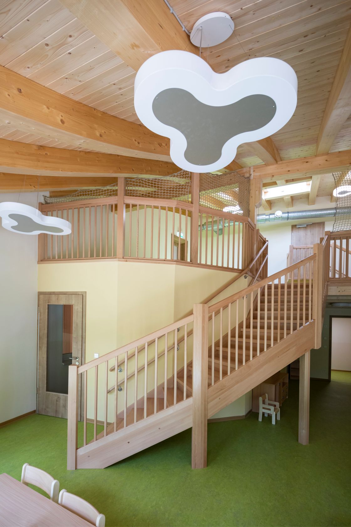 Restaurant des Kindergartens Am Wald- Klinikum mit Holztreppe und Wolkenlampen Innenraum mit einer Holztreppe, die nach oben führt. Die Treppe hat ein Geländer aus Holz. An der Decke sind große Holzbalken und Lampen in Form von Wolken. Der Boden ist grün. Im Hintergrund sieht man eine Tür und einen Bereich mit hellen Wänden. Der Raum wirkt offen und freundlich