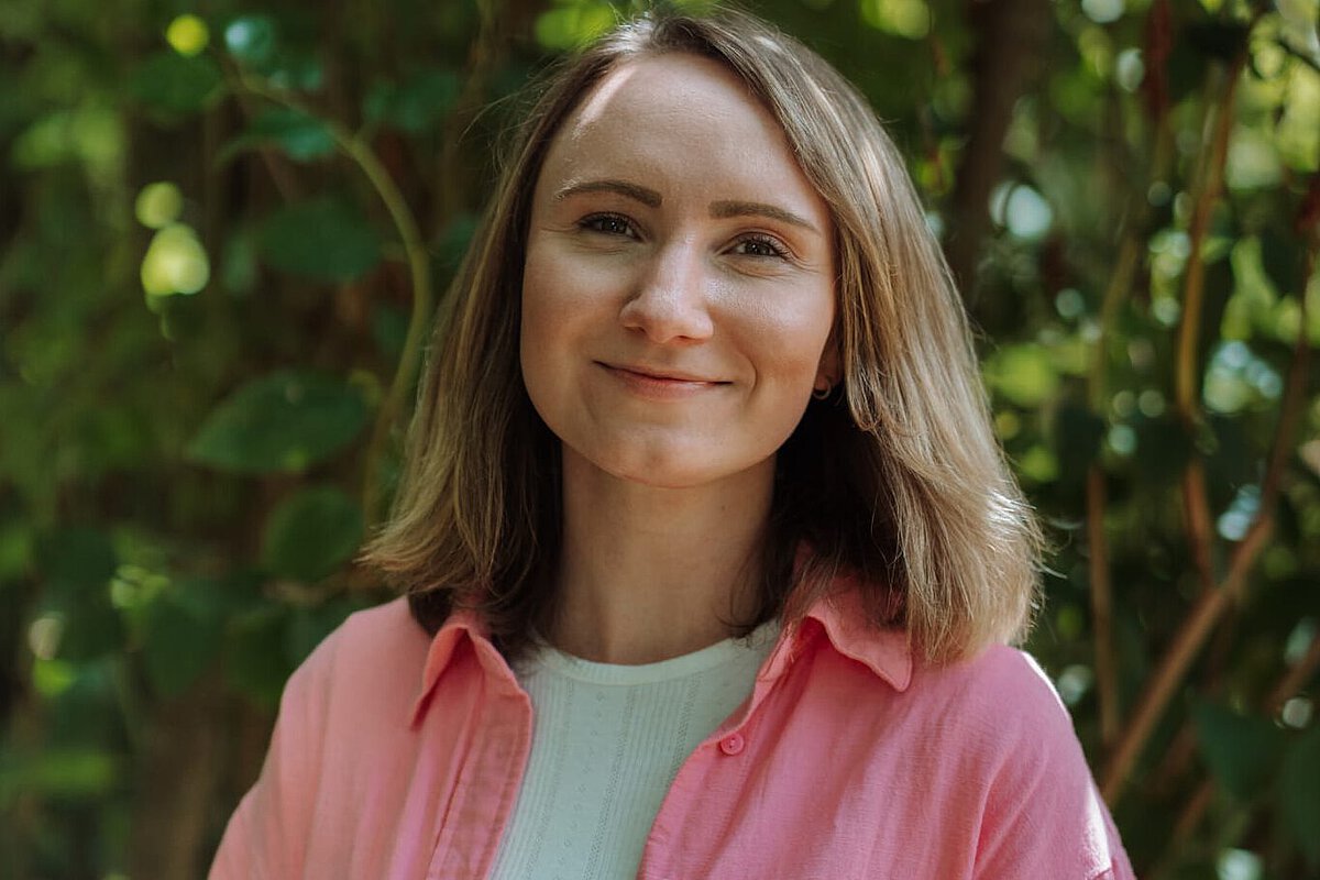 Interviewpartnerin Constanze Winder aus dem Montessori Kindergarten Tümplingstraße Constanze Winter aus dem Kindergarten Tümplingstraße steht vor einem grünen Hintergrund mit weißem Shirt und pinker Jacke