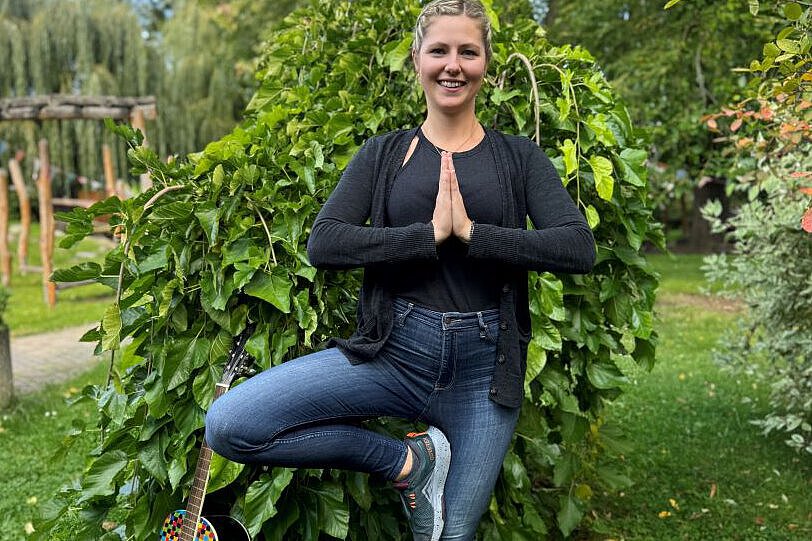 Interview mit Erzieherin Laura Stegmann – Kreativität und Ruhe im Kita-Alltag Erzieherin Laura Stegmann steht im Freien vor einem grünen Busch, zeigt eine Yoga-Pose und trägt eine schwarze Jacke mit Jeans. Neben ihr lehnt eine bunt bemalte Gitarre – ein Symbol für kreative pädagogische Arbeit.
