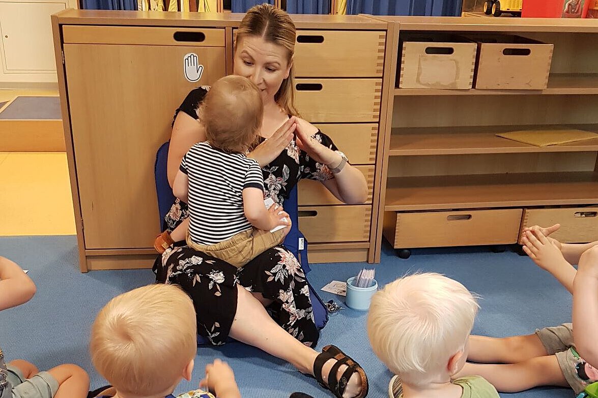 Pädagogische Fachkraft der Kita Hölderlinstraße mit Kleinkindern Person sitzt mit mehreren Kleinkindern im Kreis auf dem Boden und begleitet ein Kind durch Gesten und Interaktion.