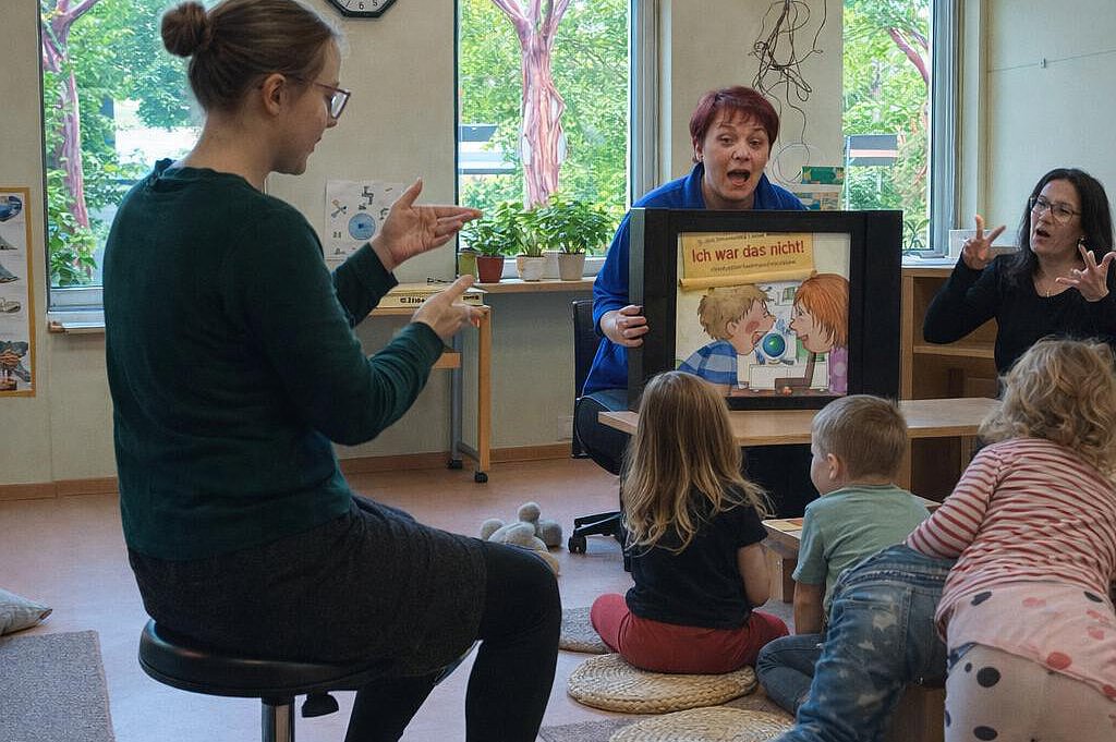 Inklusive Mediennutzung in der Kita: Vorlesen mit Gebärden und Bilderbuch Pädagogische Fachkräfte lesen Kindern in einer Kita eine Geschichte vor und unterstützen diese mit Gebärden; mehrere Kinder sitzen im Halbkreis und verfolgen die Bilder.